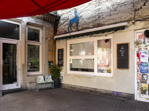 Zum Scheuen Reh Das Bild zeigt einen Außenbereich mit einer weiß gestrichenen Wand, auf der eine stilisierte Hirschfigur angebracht ist. Der Eingang in einem urbanen Straßenkontext mit Plakaten auf der Tür bietet eine einladende Atmosphäre.