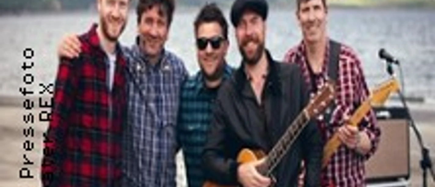 Eine fröhliche Gruppe von fünf Musikern steht am Ufer eines Sees mit Gitarren und Schlagzeug.