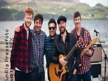 Eine fröhliche Gruppe von fünf Musikern steht am Ufer eines Sees mit Gitarren und Schlagzeug.