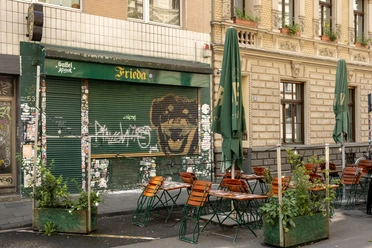 Das Bild zeigt die Fassade eines Cafés mit dem Namen "Frieda", Tische im Freien unter Sonnenschirmen platzieren, umgeben von Pflanzen. Streetart und Sticker schmücken den geschlossenen Ladenbereich.