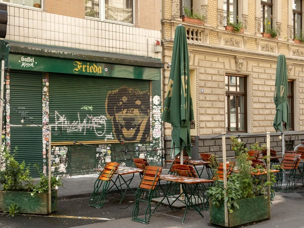 Frieda Bar Das Bild zeigt die Fassade eines Cafés mit dem Namen "Frieda", Tische im Freien unter Sonnenschirmen platzieren, umgeben von Pflanzen. Streetart und Sticker schmücken den geschlossenen Ladenbereich.