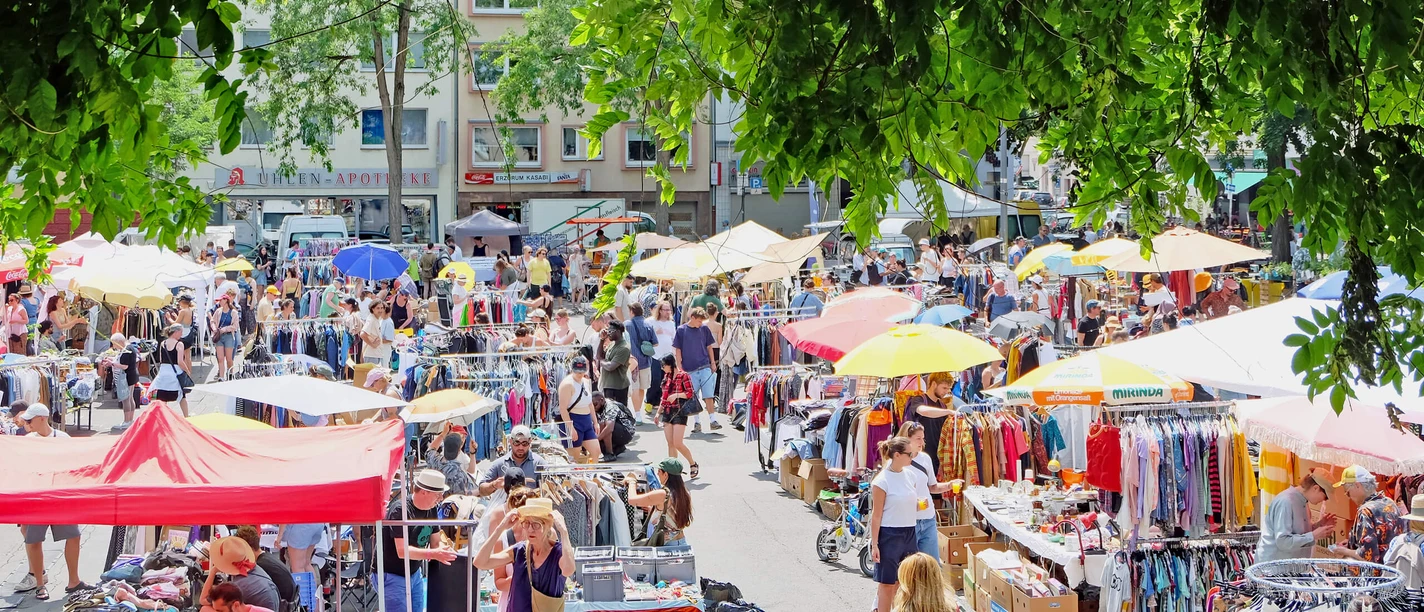 Flea market in Cologne-Nippes A lively flea market scene full of colorful stalls and busy people under shady foliage.