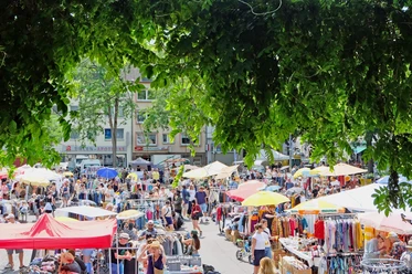 Flohmarkt in Köln-Nippes Eine lebendige Flohmarkt-Szene voller bunter Stände und emsiger Menschen unter schattenspendendem Laub.