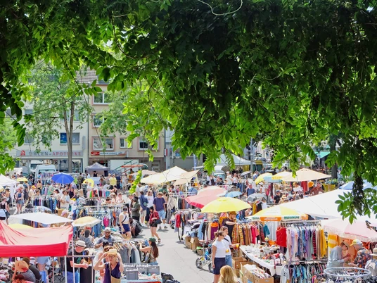 Flohmarkt in Köln-Nippes Eine lebendige Flohmarkt-Szene voller bunter Stände und emsiger Menschen unter schattenspendendem Laub.A lively flea market scene full of colorful stalls and busy people under shady foliage.