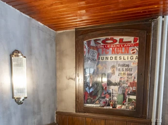 Forelle Blau Ein Holztisch mit Bank in einer mit Holz vertäfelten Ecke, darüber ein Fanposter des 1. FC Köln.A wooden table with a bench in a wood-paneled corner, with a 1. FC Köln fan poster above it.