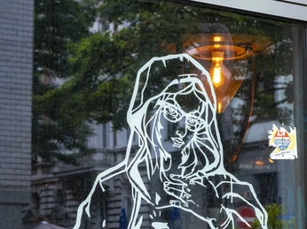 Zum Goldenen Schuss Eine Glastür mit einer weißen Umrisszeichnung eines Menschen, der eine Kamera hält. Sie reflektiert Bäume und Häuser im Hintergrund.A glass door with a white outline of a person holding a camera. It reflects trees and houses in the background.