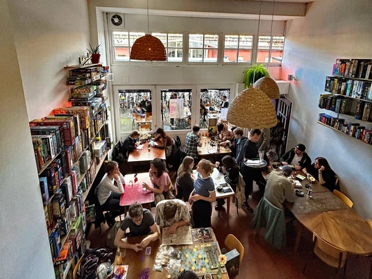 Bingo Club Cologne Ein lebendiger Raum voller Menschen, die an verschiedenen Tischen Brettspiele spielen, mit Regalen voller Spiele an den Wänden.A lively room full of people playing board games at various tables, with shelves full of games on the walls.