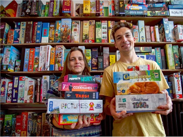 Bingo Club Köln Zwei Personen, strahlend, vor einem Spieleladenregal voller bunter Brettspiele, halten stolz ihre Favoriten in den Händen.