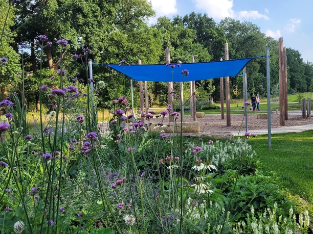 Ein Wasserspielplatz mit Blumenbeet im Vordergrund. Spielgeräte und Bäume im Hintergrund.