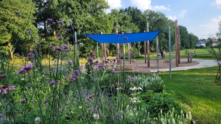 Wasserspielplatz Kurpark Levern Ein Wasserspielplatz mit Blumenbeet im Vordergrund. Spielgeräte und Bäume im Hintergrund.