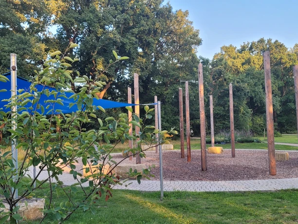 Wasserspielplatz Kurpark Levern Wasserspielplatz mit hohen Holzstämmen, Kieselbelag und blauen Sonnensegeln im Kurpark Levern.