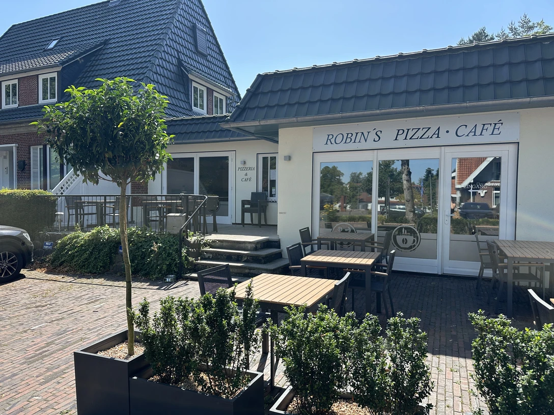 Auf der Terrasse von Robin's Pizza-Cafe Außenbereich eines Cafés mit Holztischen und Stühlen vor einem hellen Gebäude bei Sonnenschein.