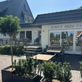 Auf der Terrasse von Robin's Pizza-Cafe Außenbereich eines Cafés mit Holztischen und Stühlen vor einem hellen Gebäude bei Sonnenschein.