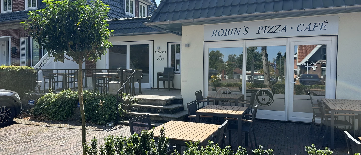 Auf der Terrasse von Robin's Pizza-Cafe Außenbereich eines Cafés mit Holztischen und Stühlen vor einem hellen Gebäude bei Sonnenschein.