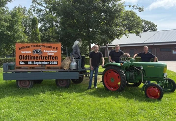 Grüne Wiese mit drei lächelnden Personen neben einem grünen Traktor und einem großen Schild.