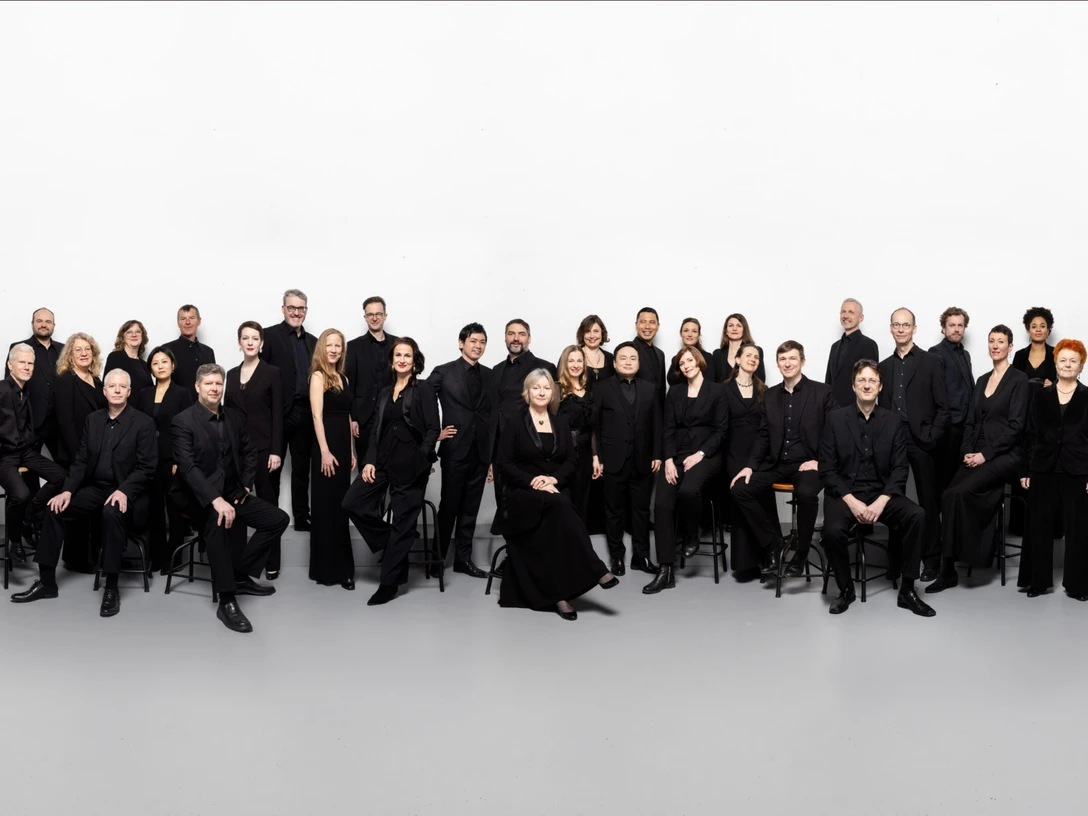 Rias Kammerchor Eine Gruppe von 28 Personen in schwarzer Kleidung posiert lächelnd in einem großen, weißen Raum.