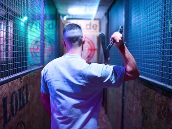 7th Space Cologne Ein Mann in einem Gang hält eine Axt, umgeben von Käfigwänden in violettem Licht.A man in a corridor holds an axe, surrounded by cage walls in violet light.
