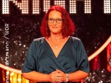 Eine Frau mit roten Haaren steht auf einer Bühne, trägt ein blaues Kleid und eine Brille. Im Hintergrund sind Lichter zu sehen.