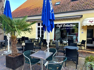 Café Schulken Außenbereich des Café Schulken mit schwarzen Sitzmöbeln, blauen Sonnenschirmen und Palme.