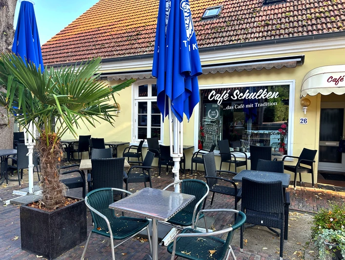 Café Schulken Außenbereich des Café Schulken mit schwarzen Sitzmöbeln, blauen Sonnenschirmen und Palme.