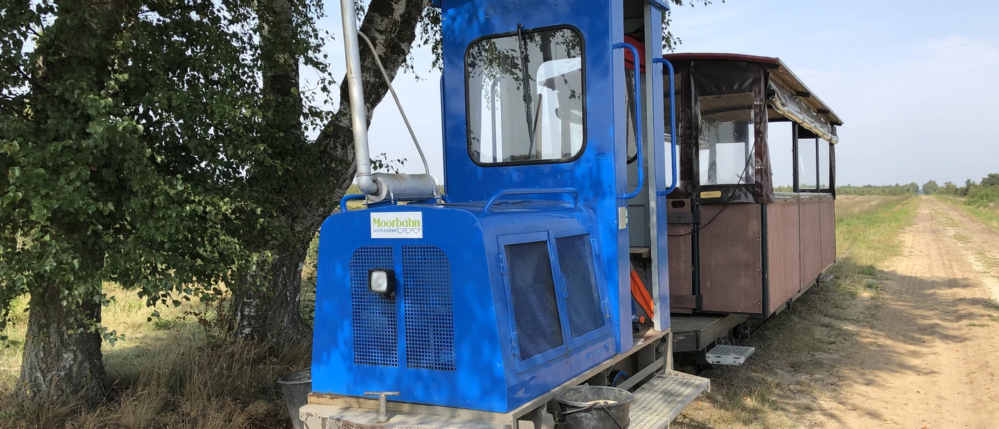 Moorbahn Uchte Eine historische Moorbahn in blau auf schmaler Schiene vor einem Baum und einem weiten, klaren Himmel.