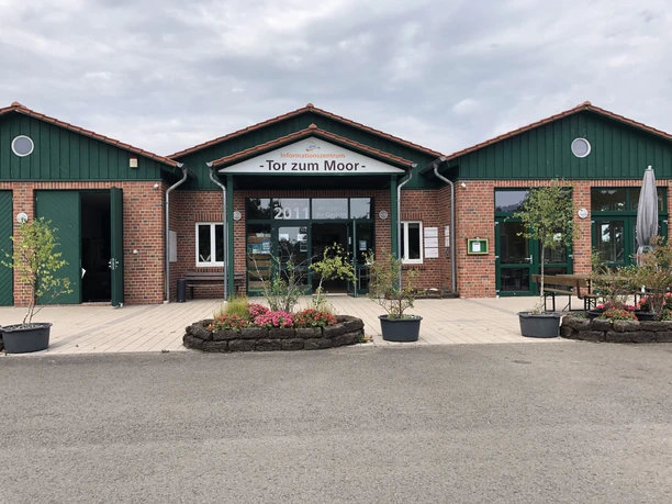 Brick building with green roof and glass fronts, surrounded by plants, with sign "Tor zum Moor".