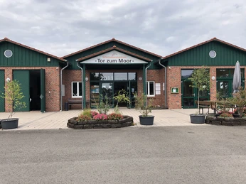 Infozentrum "Tor zum Moor" Backsteinbau mit grünem Dach und Glasfronten, umgeben von Pflanzen, mit Schild "Tor zum Moor".Brick building with green roof and glass fronts, surrounded by plants, with sign "Tor zum Moor".Murstensbygning med grønt tag og glasfacader, omgivet af planter, med skiltet "Tor zum Moor".Bakstenen gebouw met groen dak en glazen gevels, omgeven door planten, met bord "Tor zum Moor".