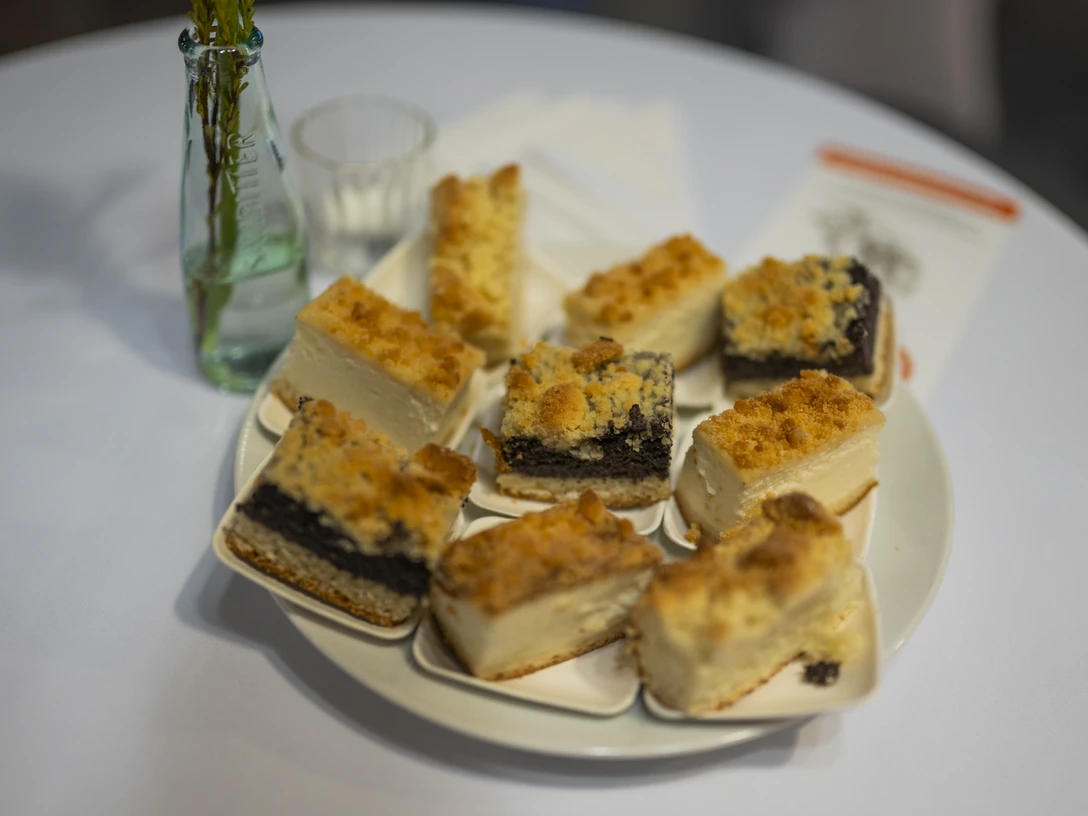 Finissage-Führung mit Kaffee und Kuchen Ein Teller mit acht Stück Mohnkuchen, umgeben von Glasgefäßen, steht auf einem weißen Tischtuch.