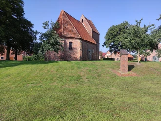 St. Antonius Kapelle, Bückelte Backsteinkapelle mit rotem Ziegeldach auf grüner Wiese, umgeben von Bäumen und Dorfhäusern bei Sonne.