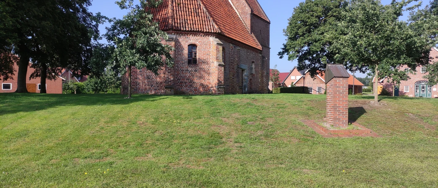 St. Antonius Kapelle, Bückelte Backsteinkapelle mit rotem Ziegeldach auf grüner Wiese, umgeben von Bäumen und Dorfhäusern bei Sonne.
