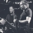 Zwei Musiker spielen Saxophon auf einer Bühne, im Hintergrund sind dunkle Lichtreflexionen zu sehen.