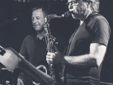 Zwei Musiker spielen Saxophon auf einer Bühne, im Hintergrund sind dunkle Lichtreflexionen zu sehen.