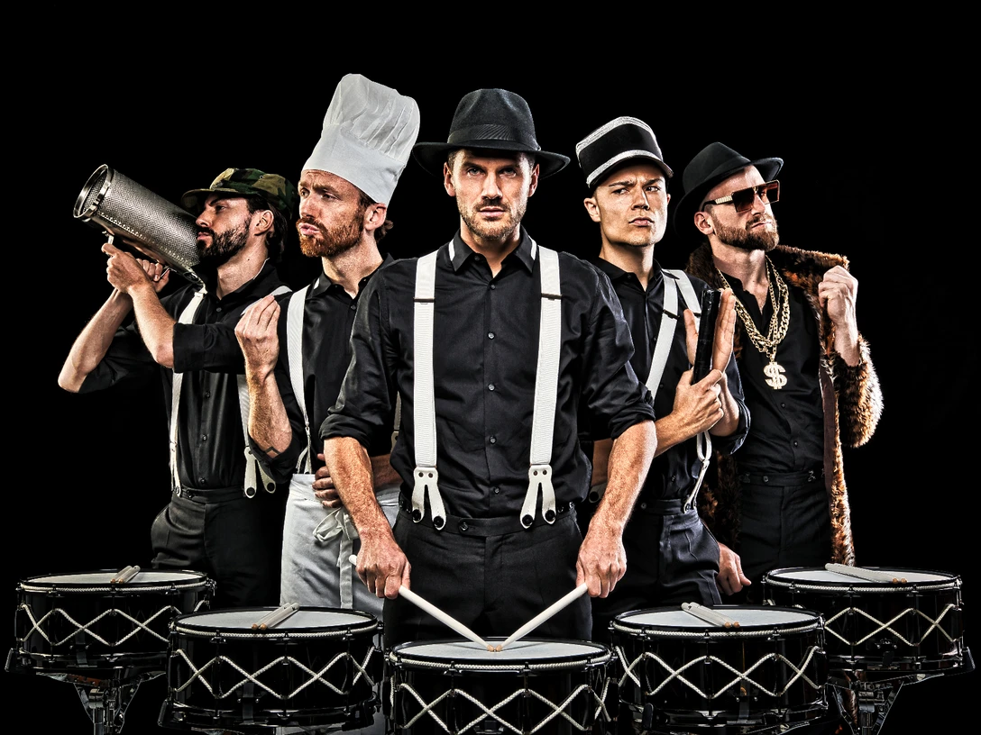 Die Schlagzeugmafia Fünf Männer in schwarzer Kleidung tragen unterschiedliche Kopfbedeckungen und spielen Trommeln.