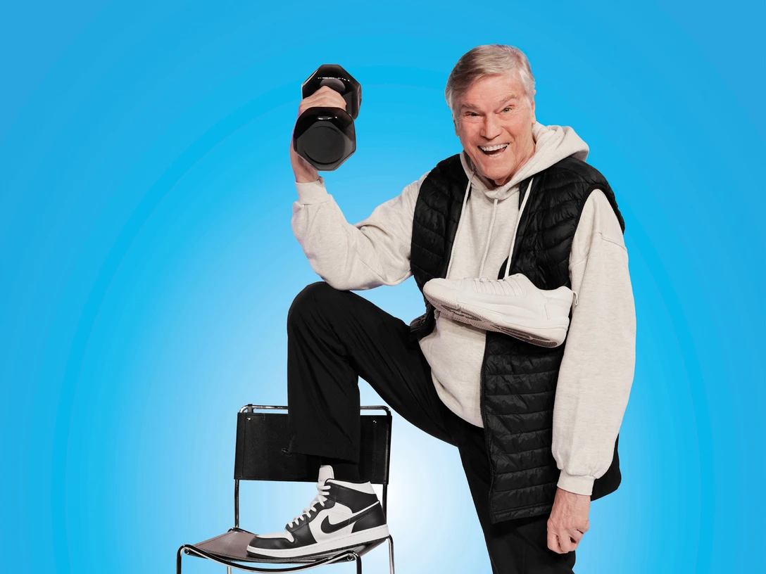 Weiße Turnschuhe "Alter schützt vor Gesundheit nicht" - Komödie von René Heinersdorff Ein älterer Herr posiert lächelnd mit Hanteln und Turnschuhen vor blauem Hintergrund auf einem Stuhl.