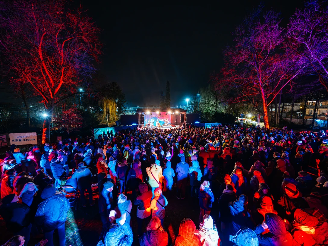 Weihnachtssingen mi Max Mutzke Eine Menschenmenge versammelt sich nachts vor einer Bühne im Freien, umgeben von bunten Lichtern.