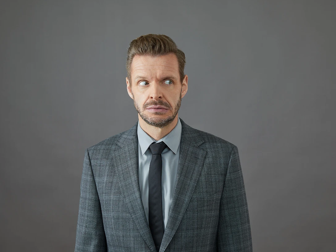 Florian Schröder Ein ernst blickender Mann im Anzug steht vor einem grauen Hintergrund und schaut zur Seite.