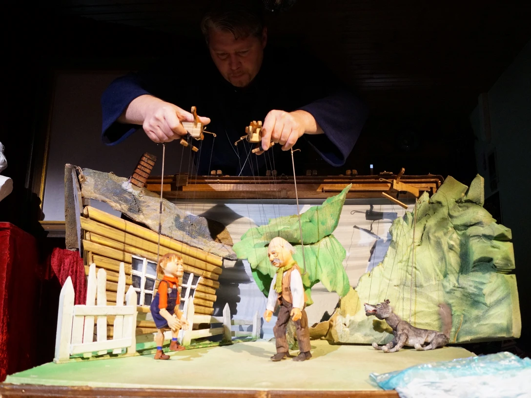 Spielszene, Peter Wolf Eine Marionettenszene von Peter und Wolf vor einem rustikalen Haus, gelenkt von Puppenspielerhänden.