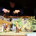 Spielszene, Peter Wolf Eine Marionettenszene von Peter und Wolf vor einem rustikalen Haus, gelenkt von Puppenspielerhänden.