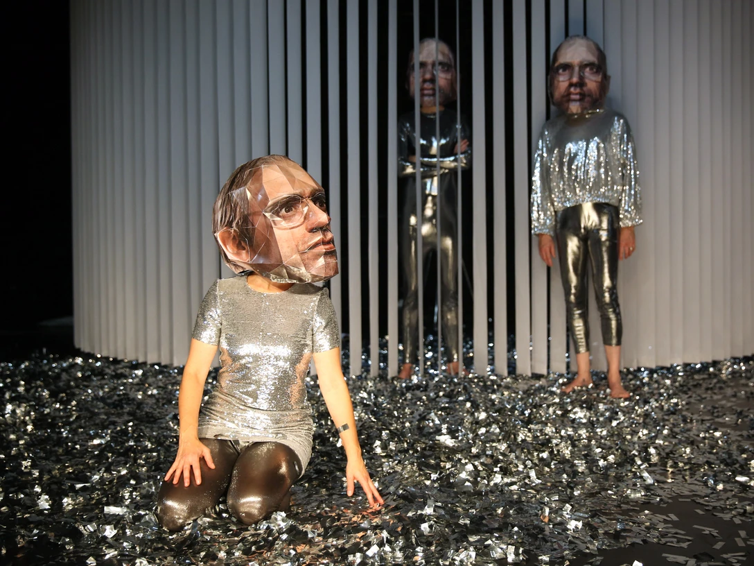 Cum-Ex Papers - Recherche zum entfesselten Finanzwesen von Helge Schmidt unter Verwendung des Materials Zwei Personen in silbernen Outfits mit großen Masken stehen und knien auf einer mit Glitzer bedeckten Bühne.