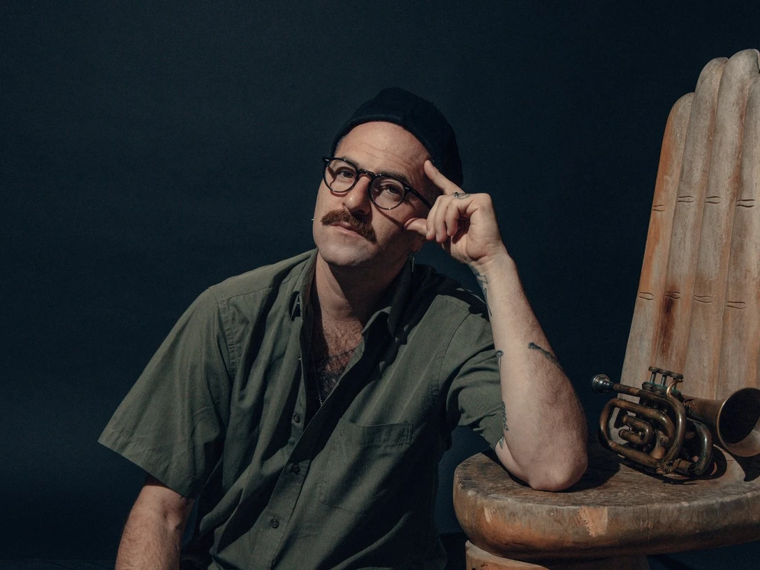 Avishai Cohen Ein Musiker mit Brille und Schnauzbart sitzt auf einem Holzstuhl, eine Trompete liegt daneben.