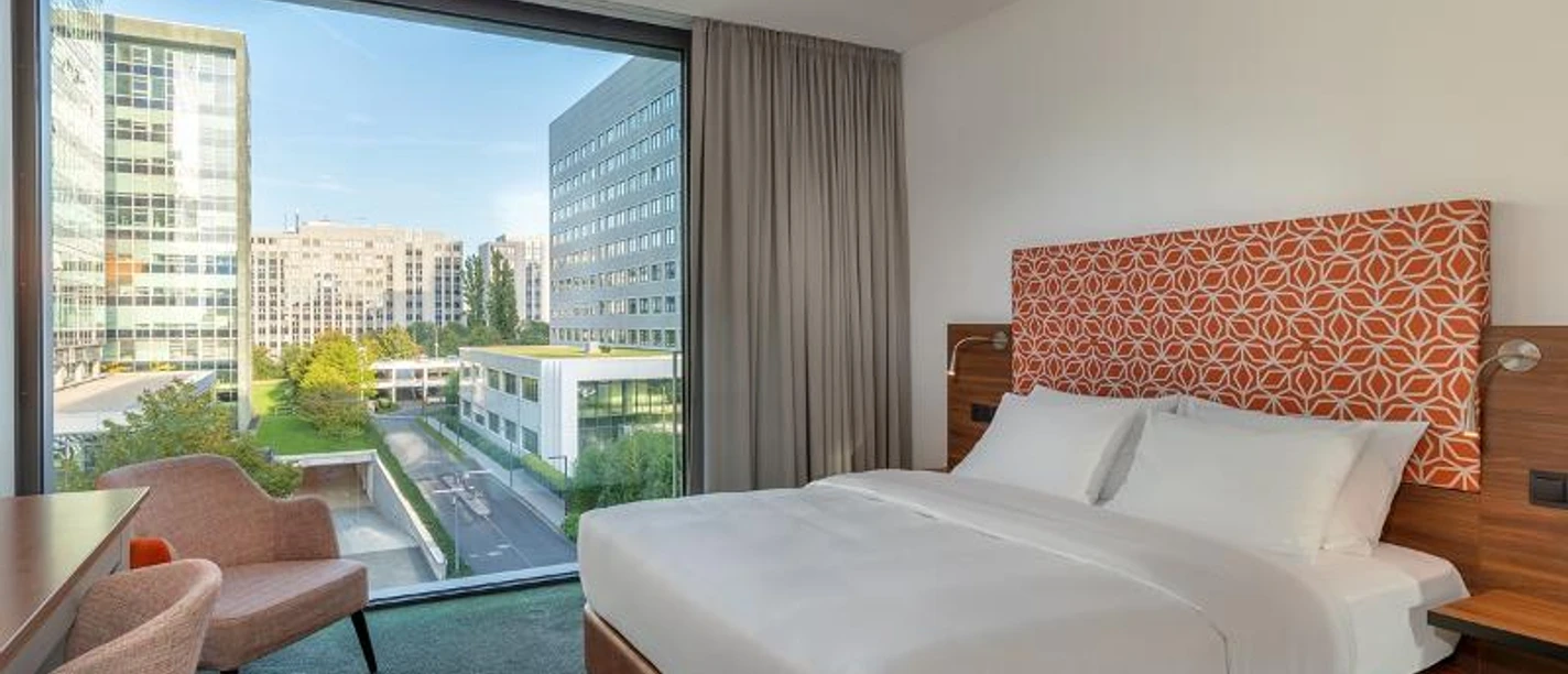 DoubleTree by Hilton Frankfurt Niederrad Ein helles Hotelzimmer mit großem Bett und Stadtblick durch das Fenster.