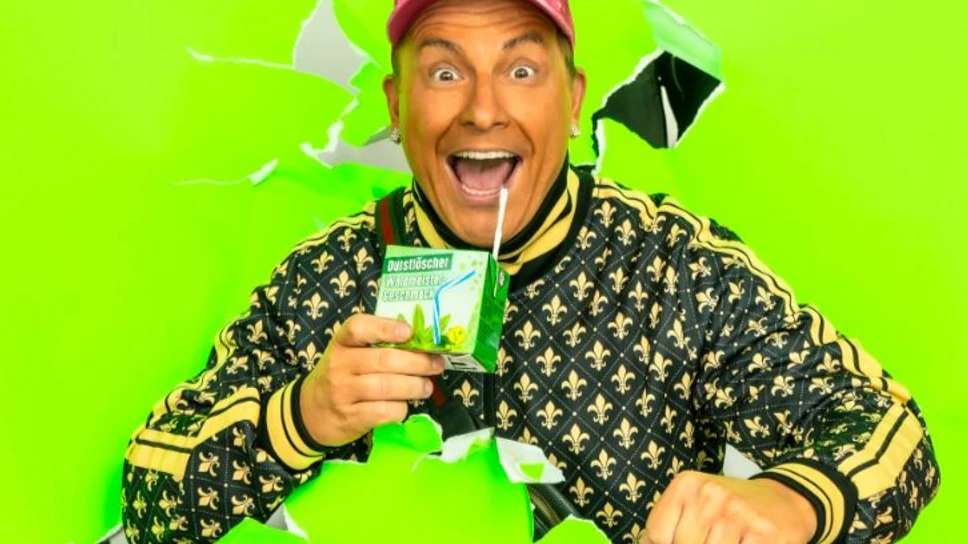 Dennis aus Hürth Ein fröhlicher Mann in auffälliger Kleidung trinkt aus einer Getränkepackung vor einem hellgrünen Hintergrund.