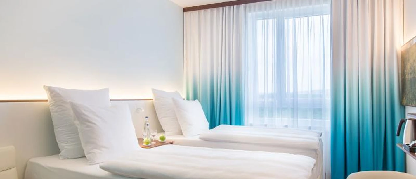 Hey Lou Hotel Frankfurt Airport Ein sauberes Hotelzimmer mit zwei Betten und blau-grünen Vorhängen.