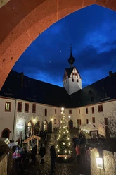 Zu sehen ist der stimmungsvoll geschmückte Hof von Schloss Rochsburg. Die Lampen am Weihnachtsbaum leuchten und es findet gerade der Adventsmarkt statt.
