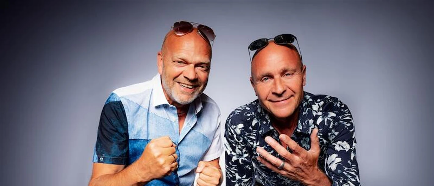 Die Feisten Zwei Männer in lässigen Sommerhemden posieren lachend vor weißem Hintergrund, Sonnenbrillen im Haar.