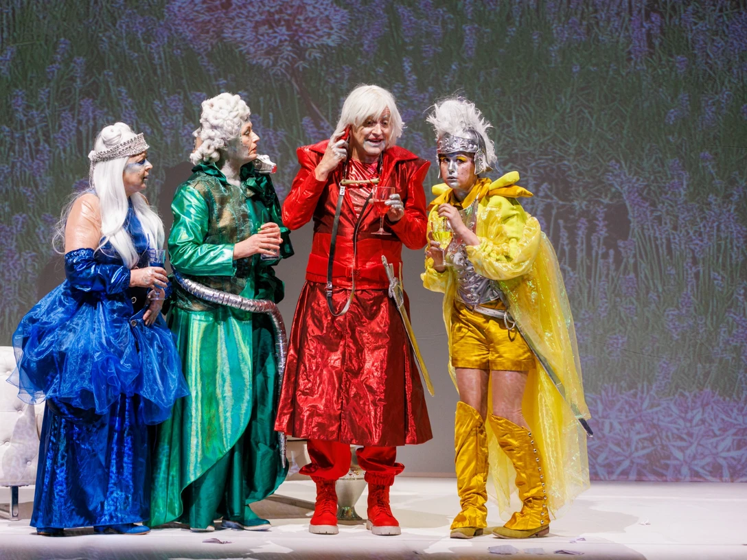 Die Guten - Schauspiel von Rebekka Kricheldorf Vier Personen in bunten, extravaganten Kostümen stehen auf einer Bühne vor einem blumigen Hintergrund.