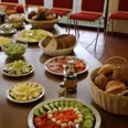 foto_montagsfruehstueck_hp.jpg Eine lange Tafel in einem Esszimmer mit umfangreichem Frühstücksbuffet, inkl. Brötchen, Obst und Gemüse.