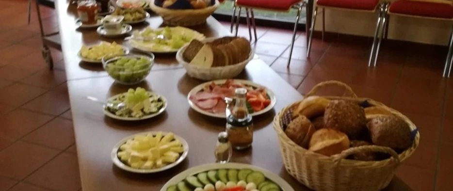 foto_montagsfruehstueck_hp.jpg Eine lange Tafel in einem Esszimmer mit umfangreichem Frühstücksbuffet, inkl. Brötchen, Obst und Gemüse.