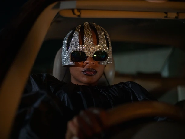 ON BECOMING A GUINEA FOWL Eine Person mit dekorativer Kristallmaske und Sonnenbrille sitzt in einem Auto bei Nacht.