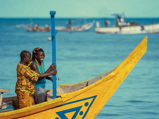 THE FISHERMAN An older and a younger man stand smiling on a yellow fishing boat with a blue mast.